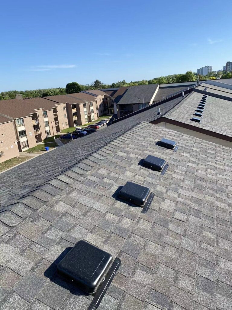 Grey asphalt shingle roof on a multi-unit residential building, fitted with a row of box vents along the ridge, overlooking nearby apartment buildings and a parking lot under a clear blue sky