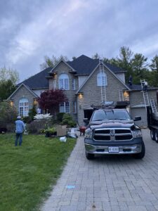 A residential driveway scene featuring a dark pickup truck parked in front of a large two-storey brick and stone home with a dark shingled roof. A person is visible near the side of the house, and mature trees and shrubs frame the property. The sky is overcast with clouds.