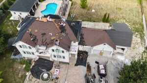 Aerial drone view of a large stone-clad estate home mid roof tear-off and reshingling, with workers, tarps, debris, shingle bundles, and an in-ground pool and patio visible in the backyard